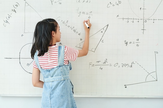 Smart schoolgirl writing on whiteboard when solving geometry task in class, view from the back