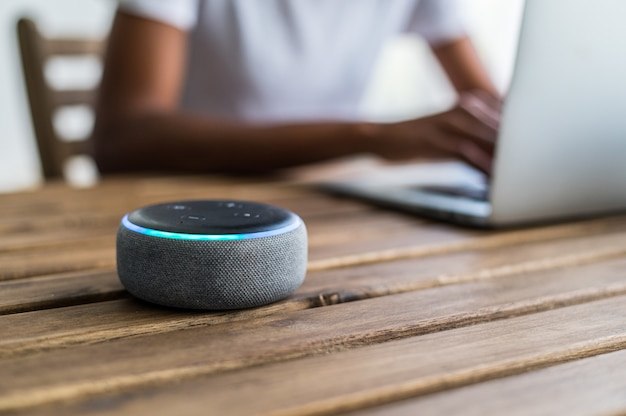 Photo smart speaker placed on table near crop black female using laptop at home