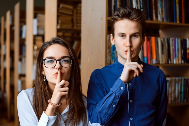 Smart student nerds at the library looking into the camera and making silence gesture