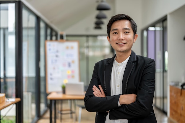 A smart and successful Asian businessman or male CEO in a formal business suit stands with his arms crossed in the office