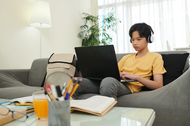 Smart teenage boy in headphone programming or playing on laptop when sitting on sofa at home