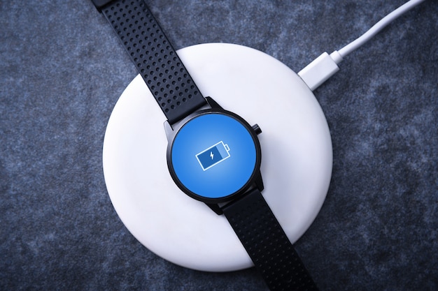 Smart watch on wireless charging with on-screen charging indicator. At the desktop, near the laptop. Top view. 