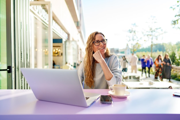 Donna astuta con gli occhiali che si siede nella caffetteria usando il computer portatile