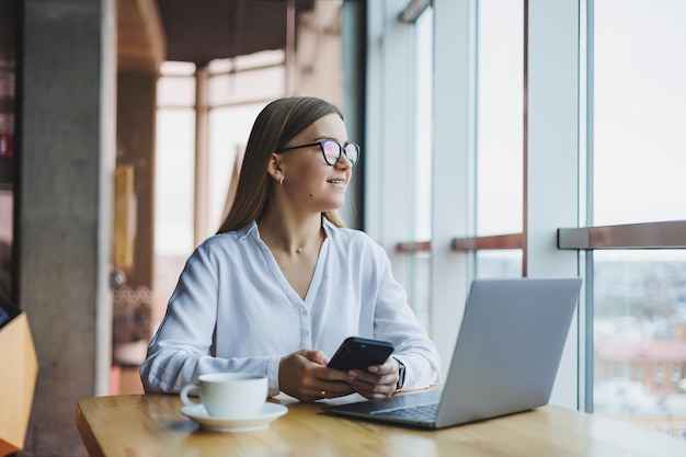 A smart woman manager with a cup of coffee has a business conversation via a smartphone to discuss a delivery order a young woman small owner sits at a table in a cafe with a laptop and calls