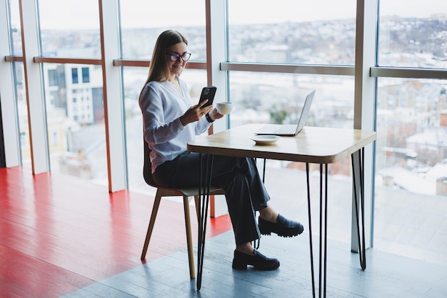 A smart woman manager with a cup of coffee has a business conversation via a smartphone to discuss a delivery order a young woman small owner sits at a table in a cafe with a laptop and calls