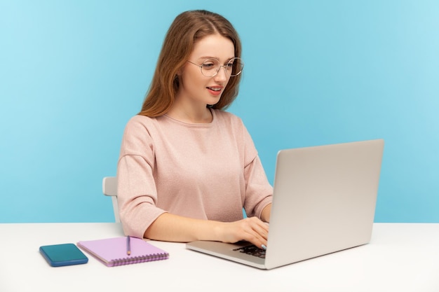 Smart woman in nerd eyeglasses typing on laptop, working with clever diligent expression in office. Student studying, doing online internet course. indoor studio shot isolated on blue background