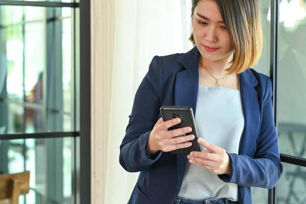 Photo smart woman in suit holding smartphone leaning against office window