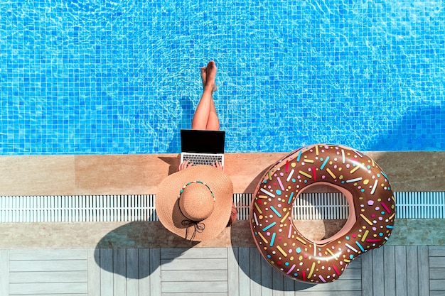 Smart woman traveler sitting with inflatable rubber circle and browsing online using computer by the swimming pool Leisure weekend and remote freelance work concept