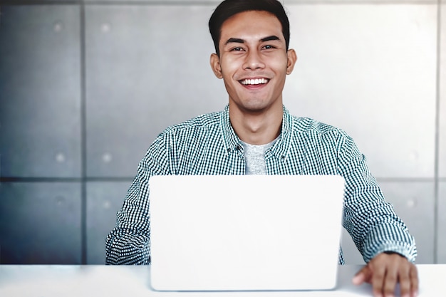 スマートの若いアジア系のビジネスマンがオフィスでラップトップコンピューターに取り組んでいます。笑顔でカメラ目線