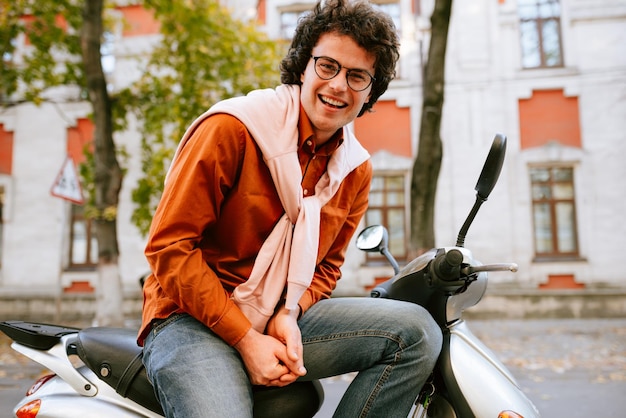 Smart young man take a break on his scooter dressed in casual outfit and transparent eyeglasses with curly hair has joyful expression