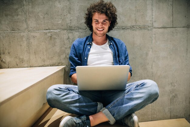 Photo smart young man with curly hair using laptop for working online surfing on internet sitting on the stairs at concrete wall handsome male reading and typing on his laptop computer in the office