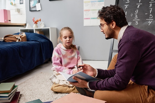 Smart young mixed race tutor in sweater sitting on carpet and holding copybook while working with li