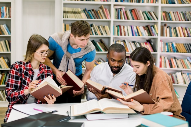 Smart giovani studenti multietnici che leggono libri al tavolo, cercano informazioni e studiano insieme nella biblioteca del college