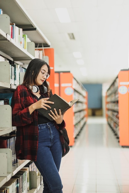 Smart young woman reading book while standing on book shelves in campus or high school library aisle