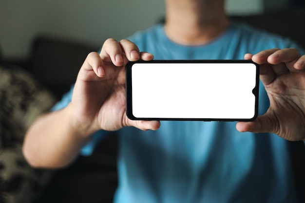 Презентация пустого экрана смартфона Молодой человек держит телефон с макетом белого пустого экрана, мужчина носит синюю футболку в гостиной дома