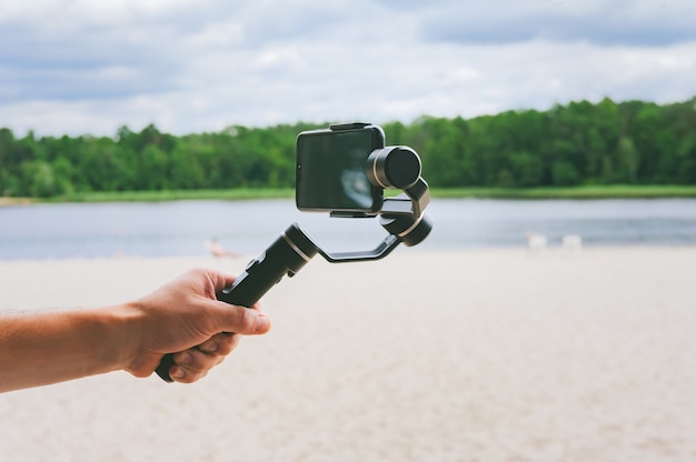 Smartphone-camerastabilisator in de hand van een man. Tegen de achtergrond van een zandstrand en de natuur met een meer.
