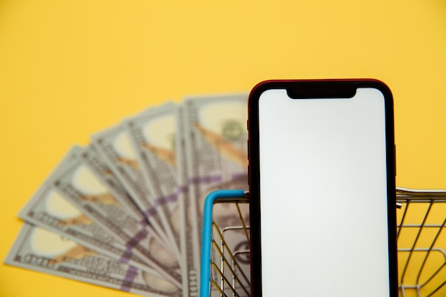 Smartphone close-up, metal market basket and banknotes of dollars on yellow