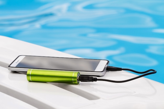 Smartphone connected to the green portable battery charger through USB cable on blue water background.