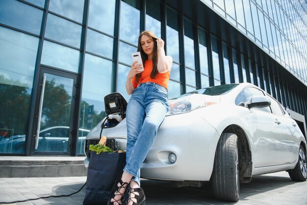 Smartphone gebruiken tijdens het wachten Vrouw op het laadstation voor elektrische auto's overdag Gloednieuw voertuig