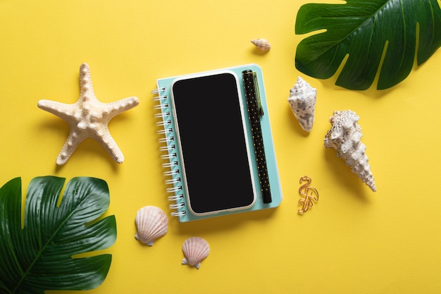 A smartphone glasses a blank notebook and Monstera Deliciosa leaves and shells on a yellow background Blogging in the summer at the workplace is a business