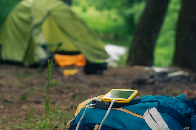 Smartphone is charged using a portable charger. Power Bank charges the phone outdoors with a backpack against the backdrop of a tent.