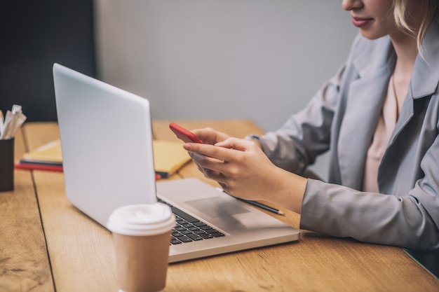 Smartphone, messaggio. mani graziose femminili che tengono smartphone sulla tastiera del computer portatile aperto in ufficio, è visibile la parte inferiore del viso della donna