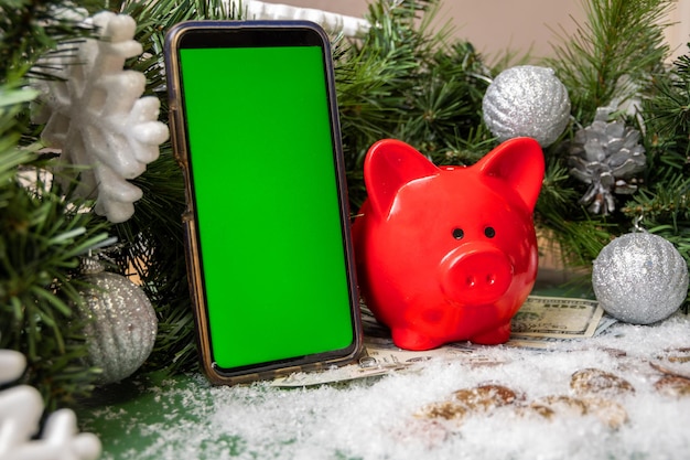 Smartphone met blanco groen scherm, rood spaarvarken op gouden munten en papiergeld bedekt met sneeuw en kerst groene sparren takken versierd met ballen. Bedrijfsconcept.