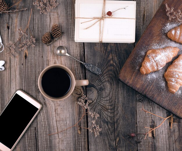 Smartphone met een leeg zwart scherm, bruine keramische kop met koffie en gebakken croissants bestrooid met poedersuiker