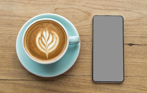 Smartphone met leeg grijs zwart scherm en warme koffie latte met latte art melkschuim in bekermok op houten bureau op bovenaanzicht. Als ontbijt In een coffeeshop in het café, tijdens zakelijk werkconcept