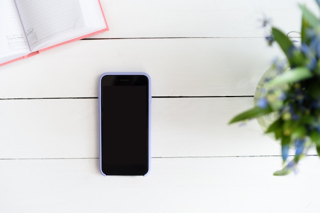 Smartphone met zwart leeg scherm. Notebook, vergrootglas en bloemen op tafel