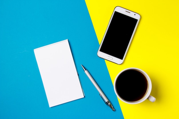 Smartphone, notepad and cup of coffee on a yellow and blue art background