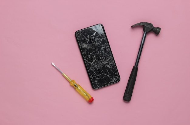 Ремонт смартфонов Смартфон с разбитым стеклянным экраном, молоток, отвертка на розовом фоне