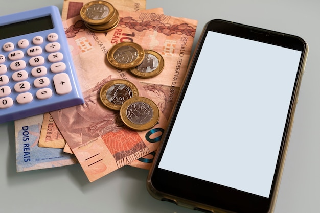 Smartphone on the table with Brazilian money coins, banknotes and calculator.