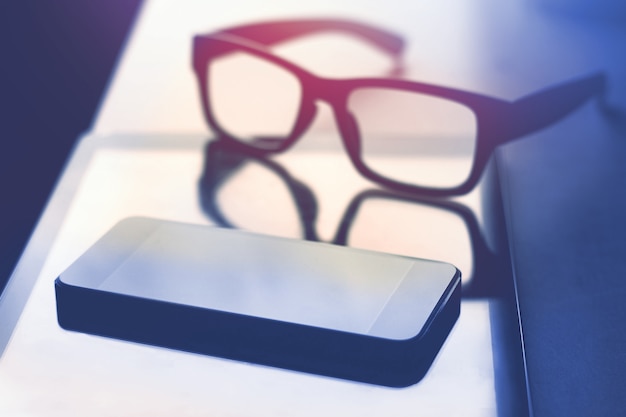 Photo smartphone tablet and eyeglasses laying on table, dark tone