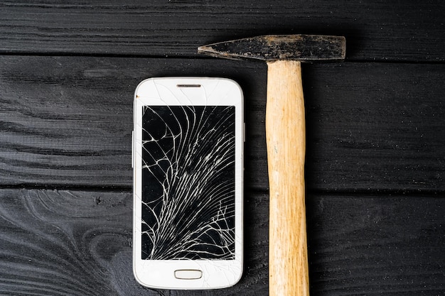 Smartphone verpletterd door hamer geïsoleerd op zwarte houten achtergrond Gebroken touchscreen van een smartphone die dicht bij de hamer op tafel ligt