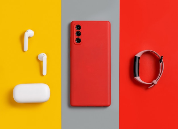 Smartphone, white wireless earphones awith the case and  smart watch top view on red, grey and yellow background