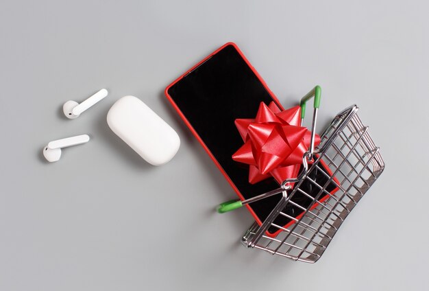 Smartphone, white wireless earphones and bow flying from the toy shopping cart top view on grey background