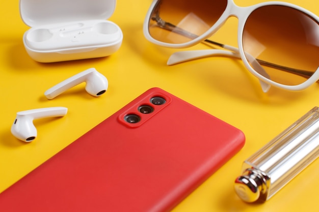 Smartphone, wireless earphones, sunglasses and lipstick on yellow background close up