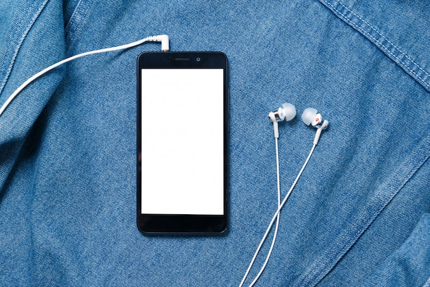 Smartphone with black case and a blank white screen and plugged in headphones ear buds on denim background