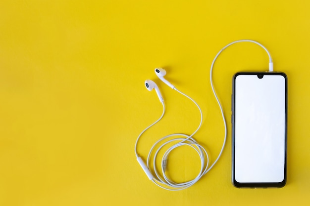 Smartphone with blank white screen connects to earphones on yellow   top view. Earphone connected to cellphone.