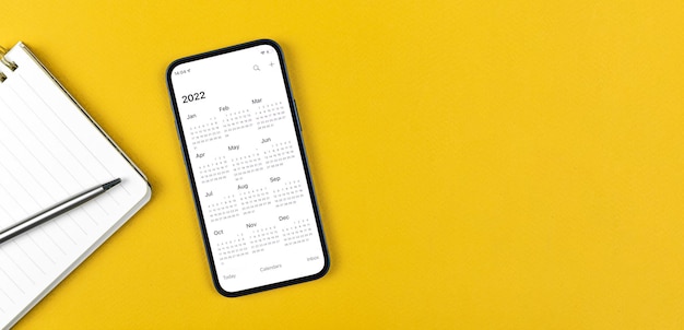 カレンダー2022のスマートフォン。ビジネスワークスペース、黄色の背景、メモ帳、銀のペン。上面写真