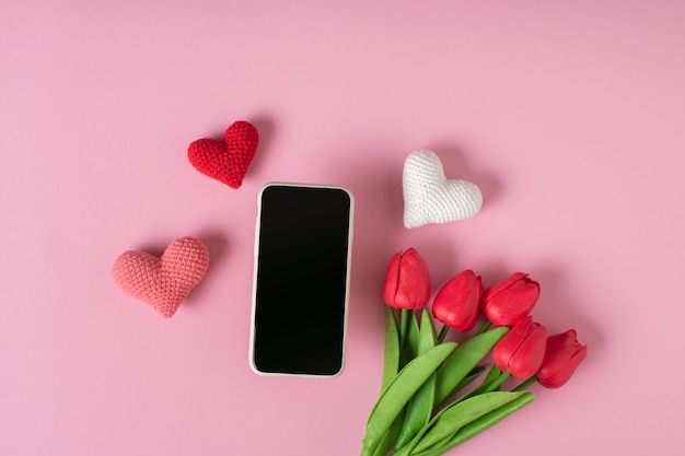 A smartphone with a clean screen with three knitted hearts and a bouquet of tulips on a pink background