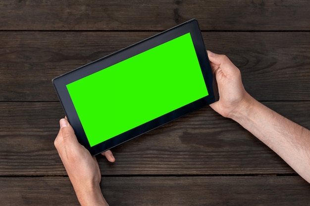 Smartphone with a green screen in hands, horizontal position, on a wooden table background.