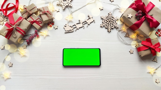 Smartphone con schermo verde sul fondo della tavola in legno bianco con i regali di natale.