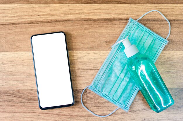 Smartphone with Protective disposable medical mask and alcohol hand sanitizer on wooden background,Top view office table