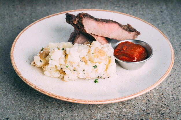 Smashed potato served with a piece of meat and red sauce on a white plate