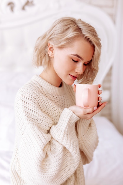 Smelling coffee. Young appealing woman wearing beige sweater feeling joyful smelling morning coffee