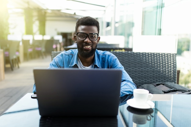Foto sorriso uomo d'affari africano seduto in caffè con in mano una tazza di caffè e utilizzando laptop