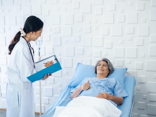 Smile Asian senior woman grey hair patient lying on bed recovering while young female doctors visit explaining the symptoms and examination notes and medical healthcare documents in hospital room