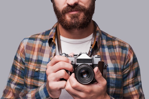 Smile to the camera! Close-up of young bearded man holding old-fashioned camera while standing against grey background